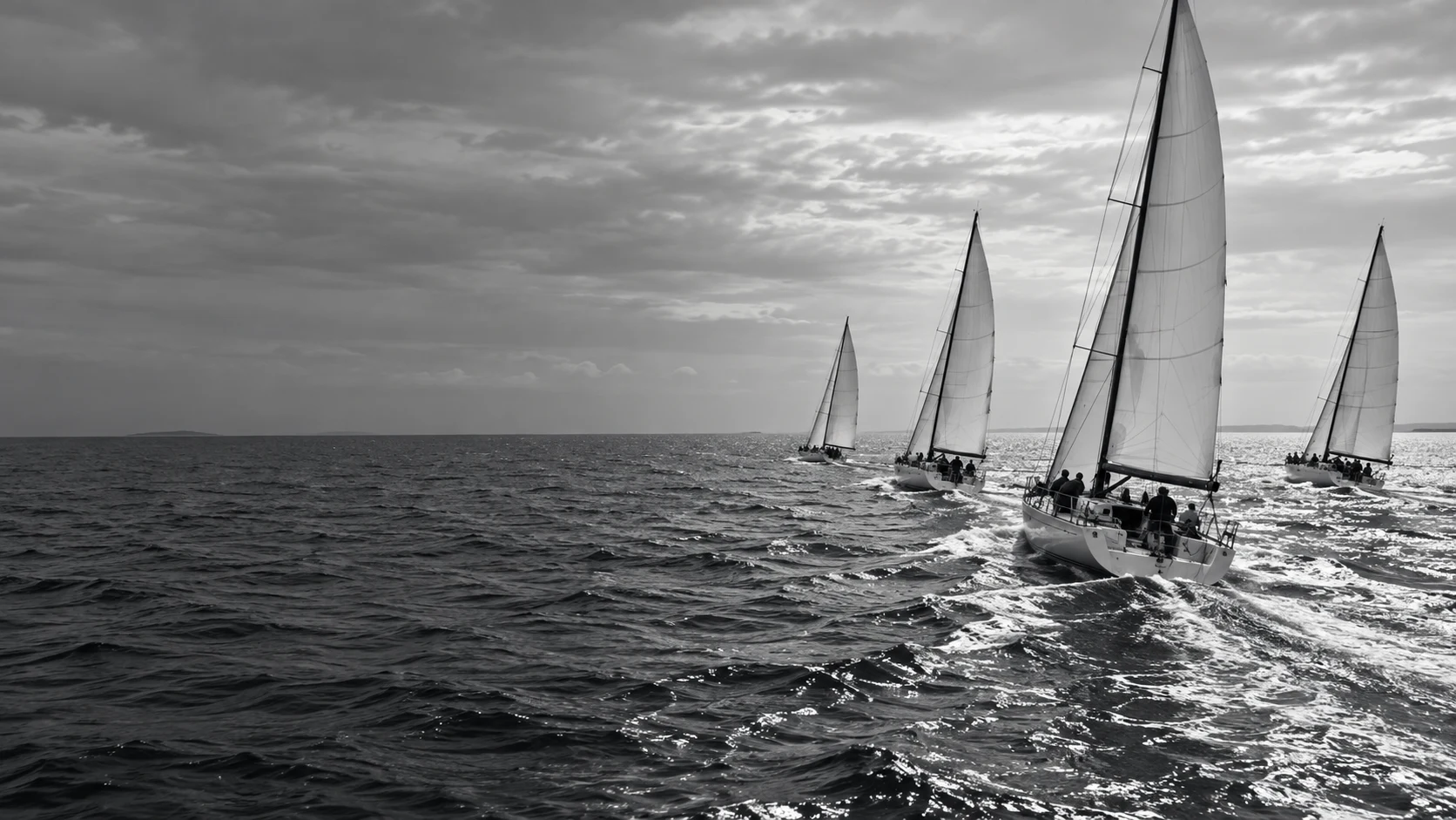 Racing sailboats advancing toward an open horizon, viewed from behind in black and white.